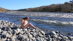 A person sits naked on river-side rocks while holding a crumpled white sheet.