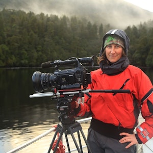 Alex Monteith is dressed in outdoors clothing standing near a large camera on a rig, they are standing next to a lake by a misty forest looking happy