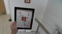 A person looks at a fire alarm switch through the lens of an iPad. Lines and geometric shapes are digitally drawn out from the switch.