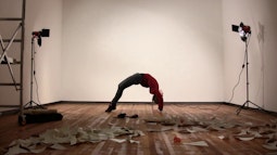 In a white walled gallery a person does a bridge pose, there aer two spotlights on either side of them. Paper is scattered on the floor.