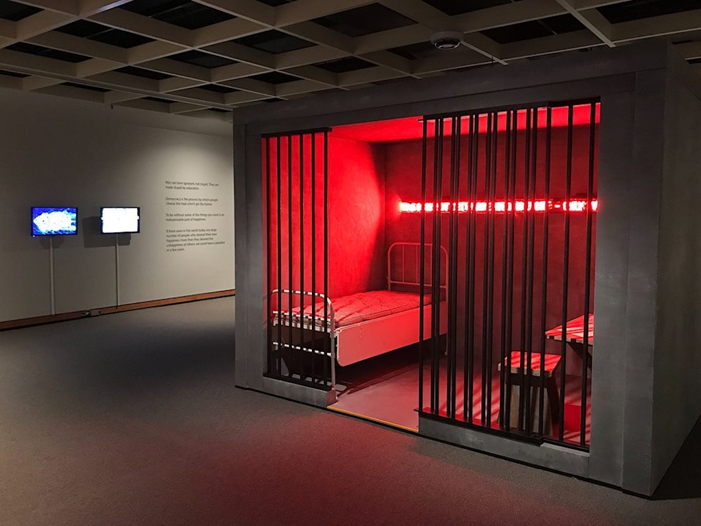 A fake prison cell iis lit with red light inside of a gallery