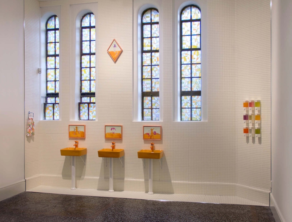 Three orange sinks are installed below stained glass windows, above each orange sink is a framed painting of the sink.