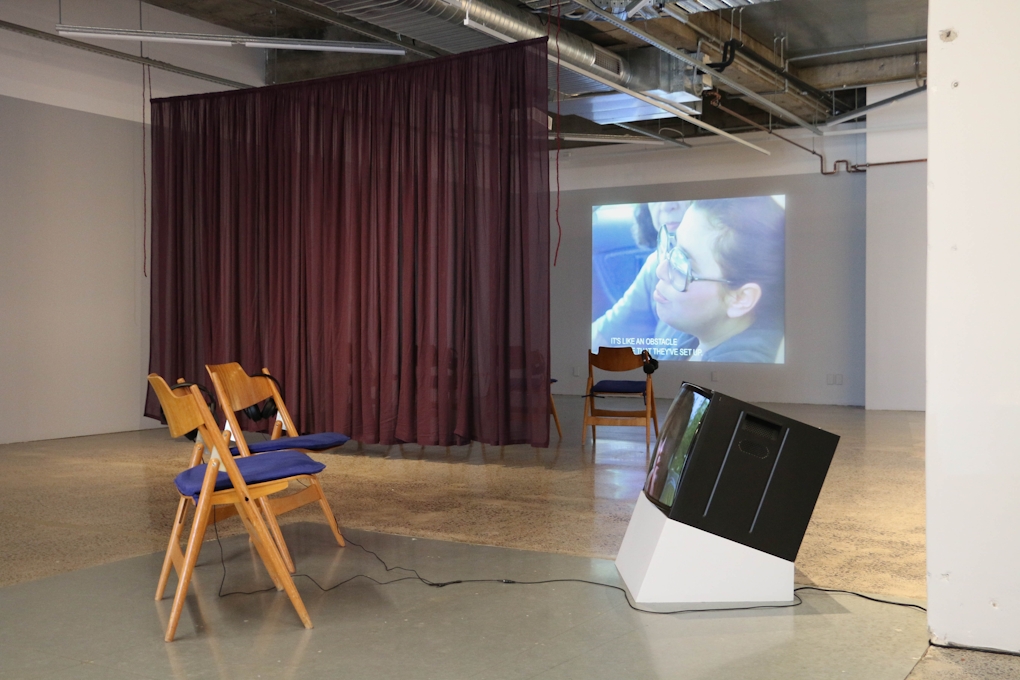 A gallery space is bisected by a sheer purple curtain. In the foreground, two chairs face a TV monitor. A film is projected onto the back wall.