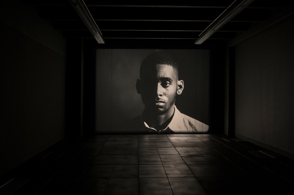 In a dark gallery a shadowy man is shown on screen looking sombre