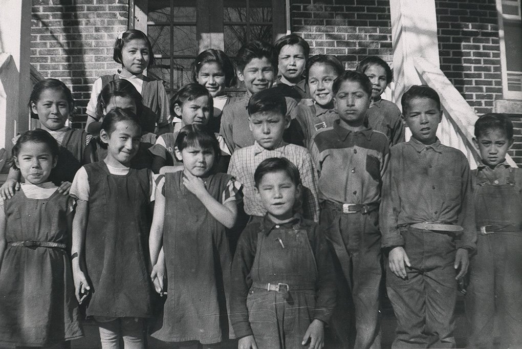 A group of First Nations children are assembled on the steps of a brick building.