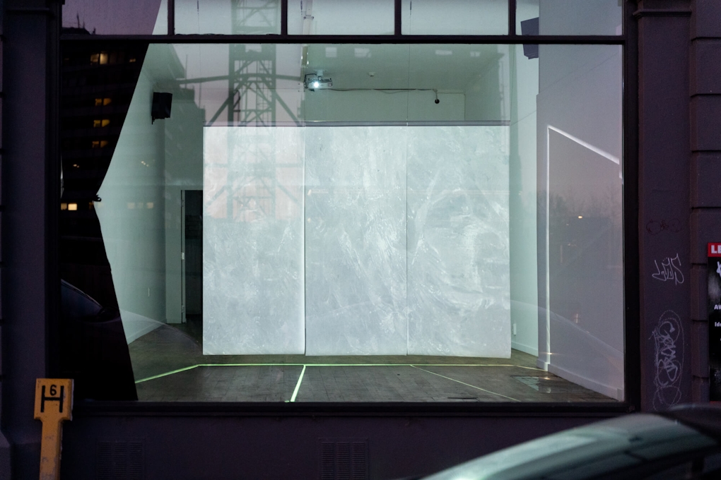 External view into a gallery window at night. A large screen inside projects an image of ice.