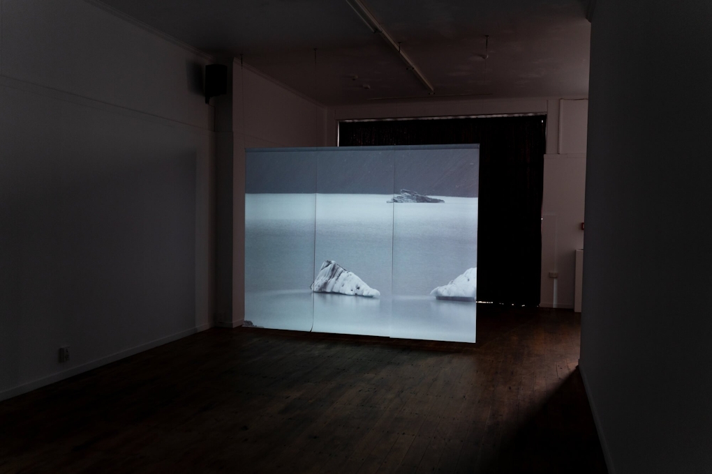 Installation view of a darkened gallery space. A large screen projects an image of icebergs in a body of water.