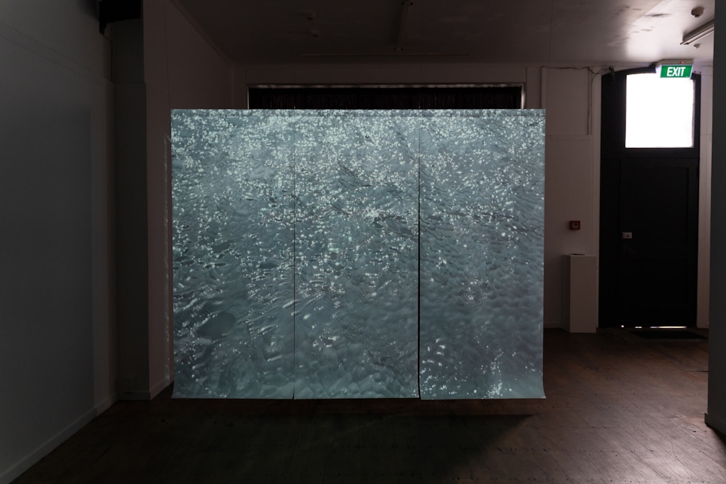 Installation view of a darkened gallery space. A large screen projects an image of water rippling.