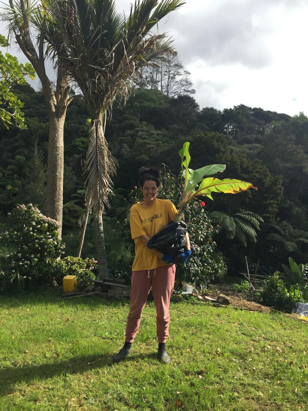 Ahilapalapa stands outside, holding a large plant with plastic over it’s roots, smiling. They are wearing a yellow t-shirt which reads “PASIFIKA”, red pants and gumboots.