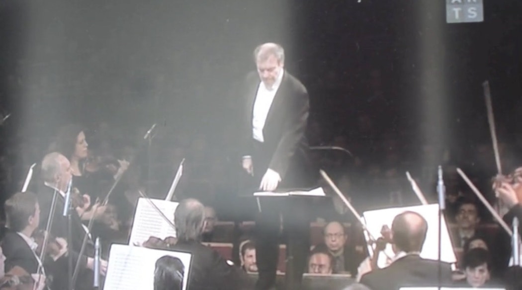 A conductor stands in front of an orchestra wearing a suit