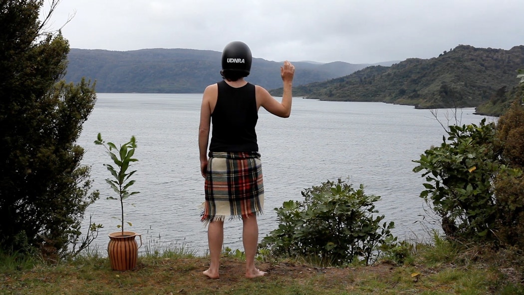 A person stands on the shoreline above a harbour. At their left is a potted plant, beside that is a naturally planted scrub bush. The person wear a tartan blanket, a black singlet and a black motorcycle crash helmet with the capital letters UDNRA printed on the back. Their right arm is raised as if in greeting or solidarity