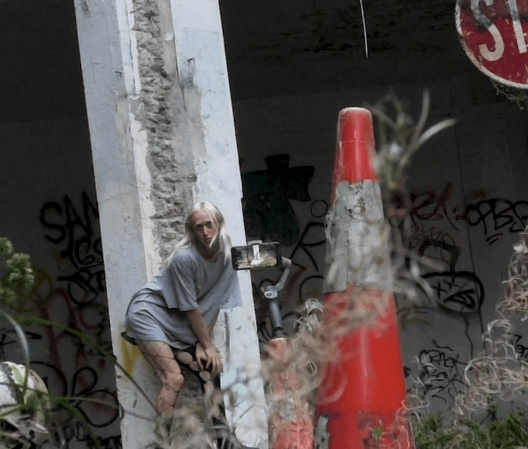 The artist leans against a tall concrete poll whilst a phone on a tripod is filming them. They are in an abandoned car park with wet concrete and weeds.