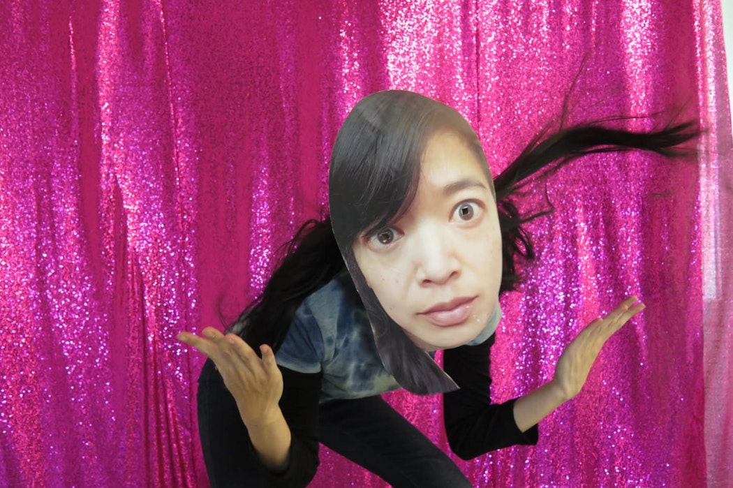 A person throws their hands up as if they are saying "what?" wearing a paper mask of Li Ming's face in front of a glitter fuschia pink curtain