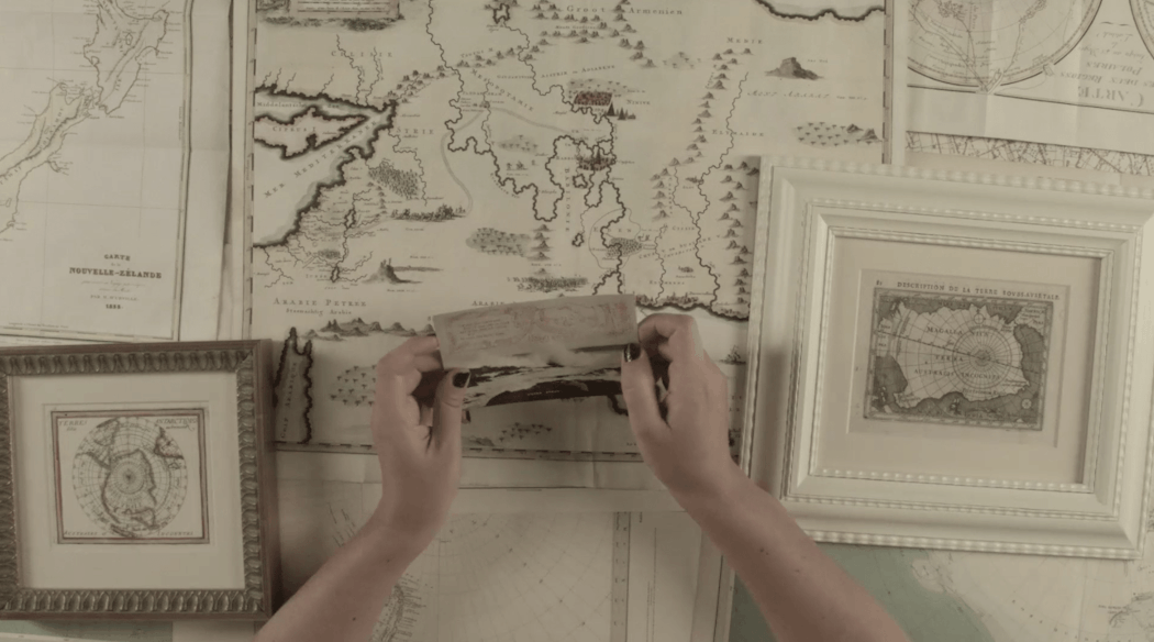 Upon a table covered with many maps, Ruth holds a postcard with two hands