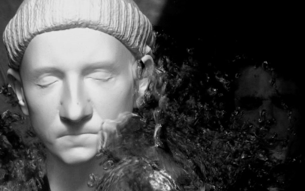 A computer generated model of the artists face is being splashed by water. The face has eyes closed and wears a beanie. It has been rendered white.