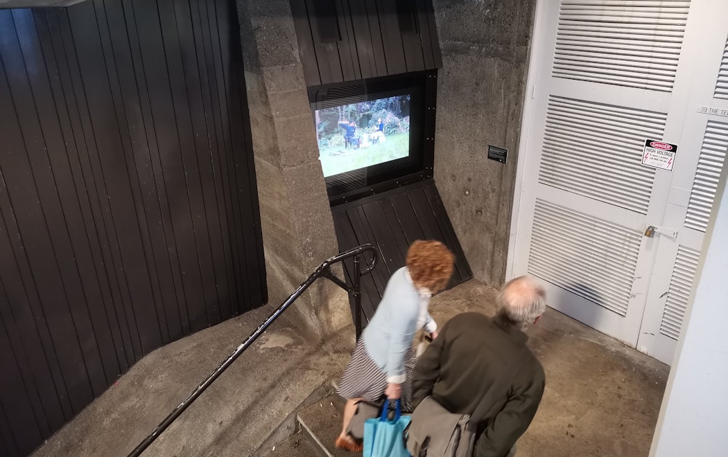 Two people are walking up the steps of Mason Lane, a central city laneway between The Terrace and Lambton Quay. They are walking past Masons Screen which is displaying a video work by Arapeta Ashton and Wai Ching Chan.