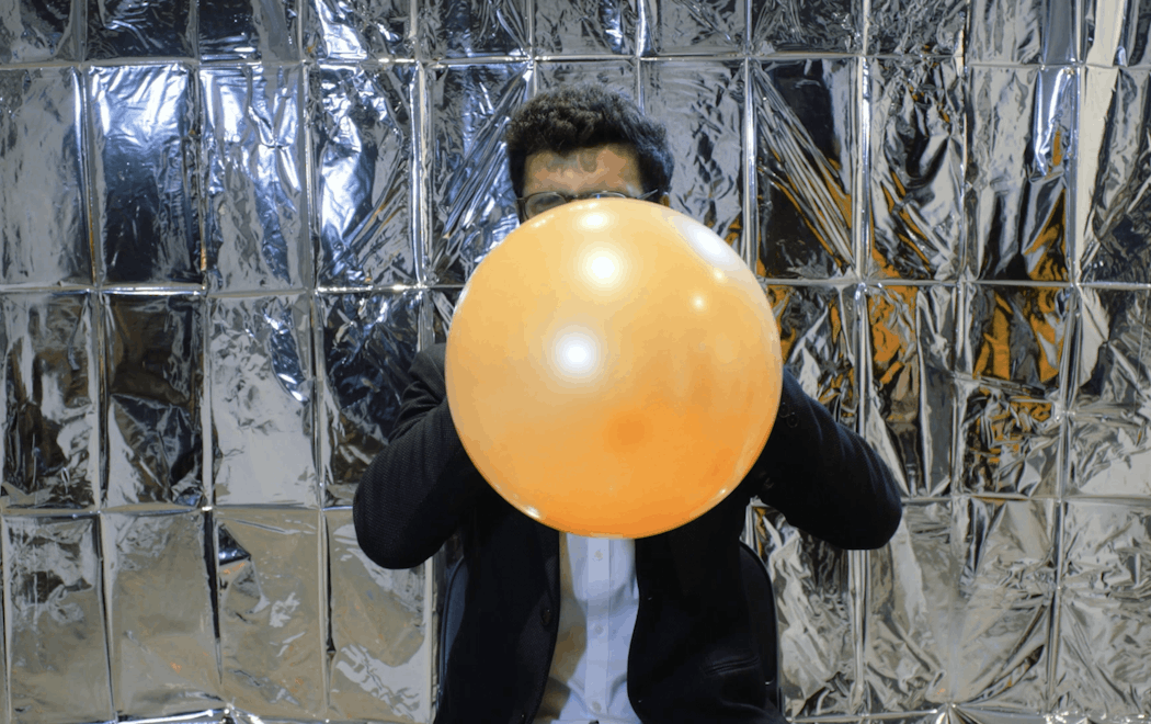A person's face is obscured by a balloon which they are inflating close to bursting