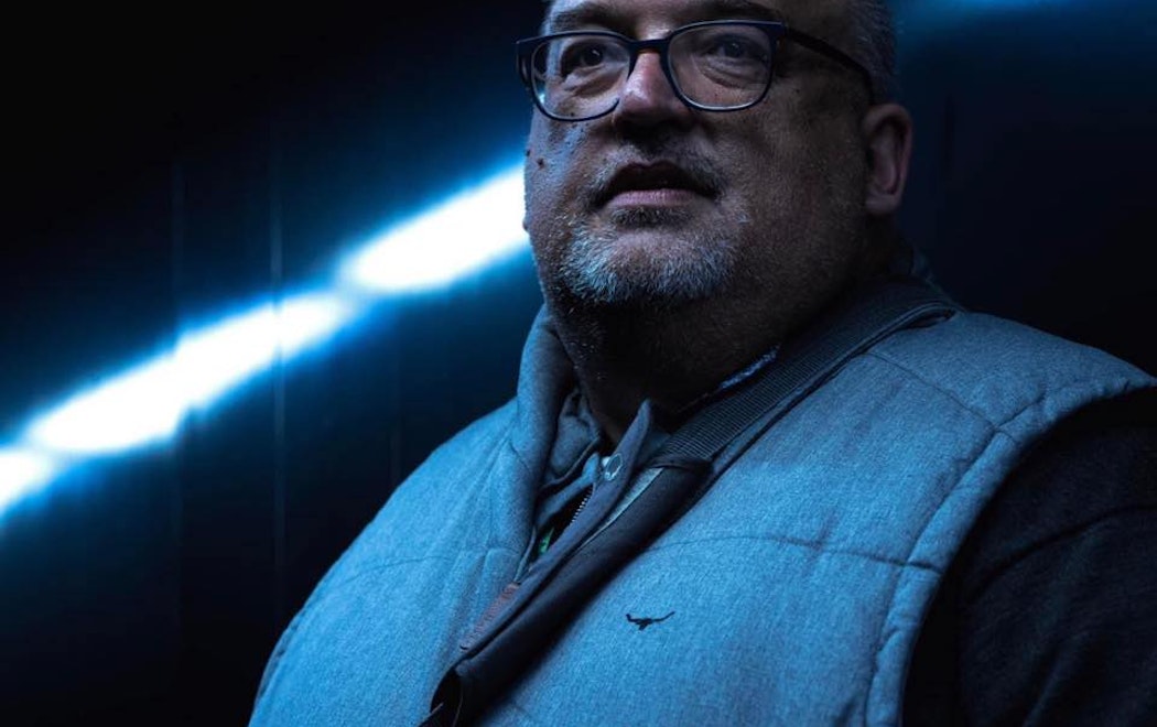 Image of a man with glasses and facial hair, wearing a puffer vest against a dark background