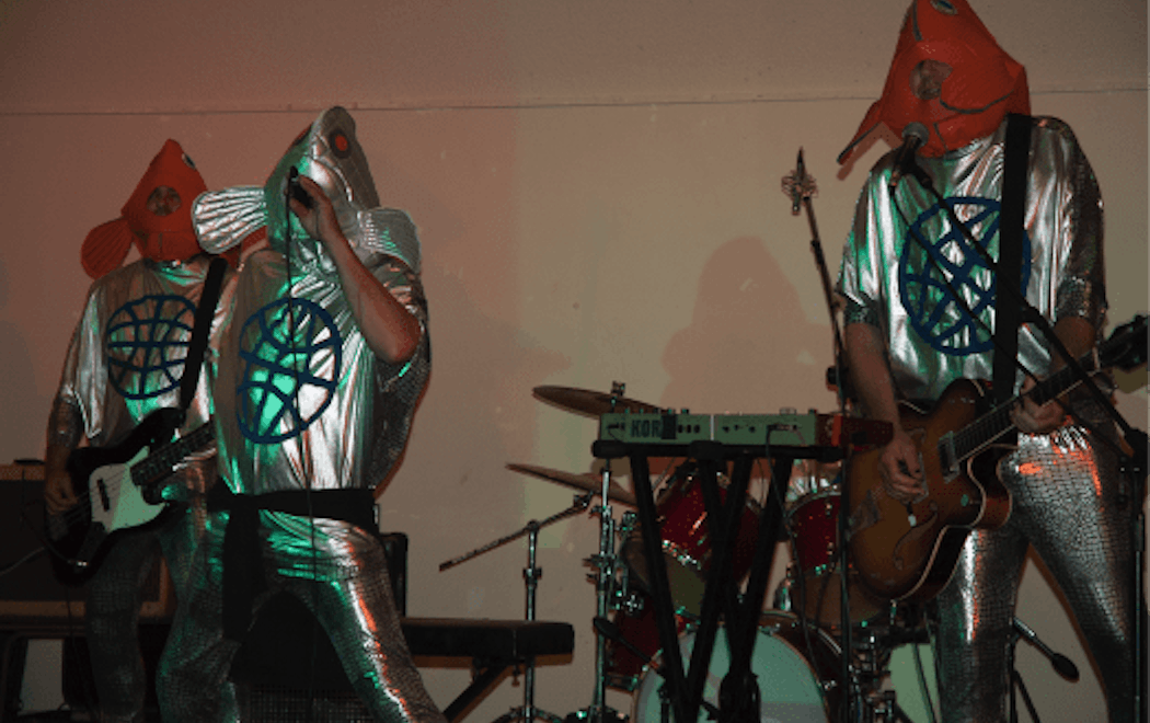 Three people perform music wearing shiny silver costumes and fish masks