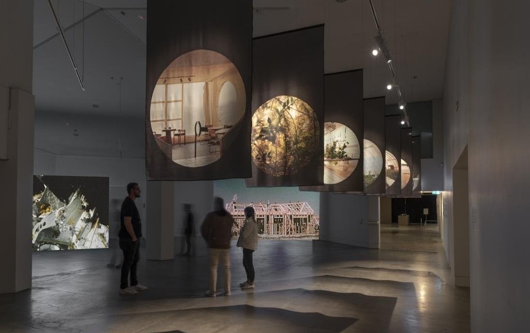 Three people stand in a gallery which displays two large screens playing video works as well as eihgt large scale photographic prints on fabric which are hung above the audiences head from the ceiling.