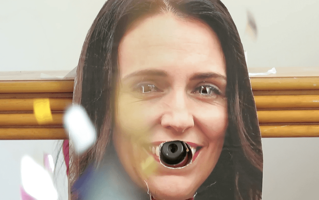 A cardboard cut-out of Jacinda Ardern has had its eyes and mouth cut out. A confetti canister is poking out of her mouth, exploding glitter