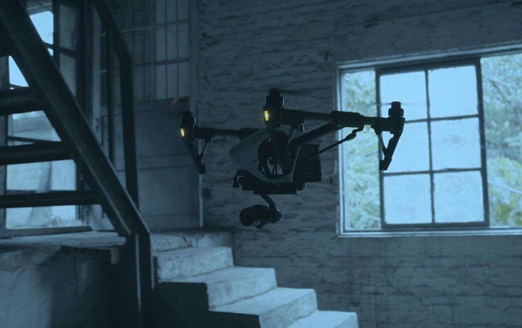 A drone flies through the inside of a building which has concrete steps and a window outside to greenery. The colour of the image is blueish grey as if its dawn, dusk or a very cloudy day