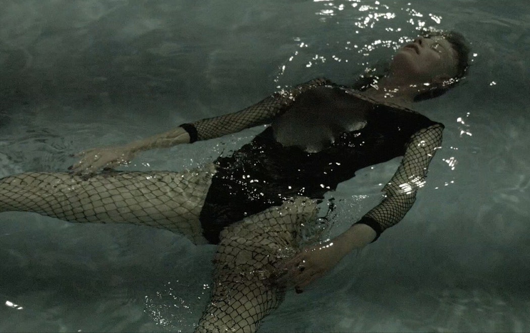 A person wearing a black swimsuit and full body fishnet is floating on their back in a swimming pool. Their head is back, floating, and eyes are closed.  they have black nail polish on. Their body is submerged apart from their chest which is slightly above water.