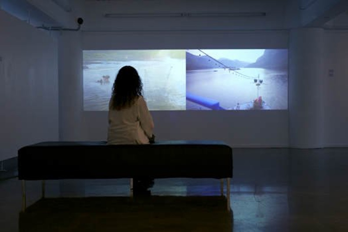 A person sits in a gallery watching a two channel video