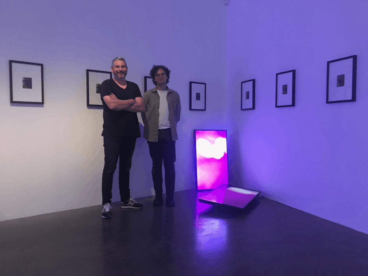Steve and Christian stand in a gallery which displays small photographs in large frames neatly on the walls, two tvs rest against the wall and the floor of the gallery displaying a bright, pink image