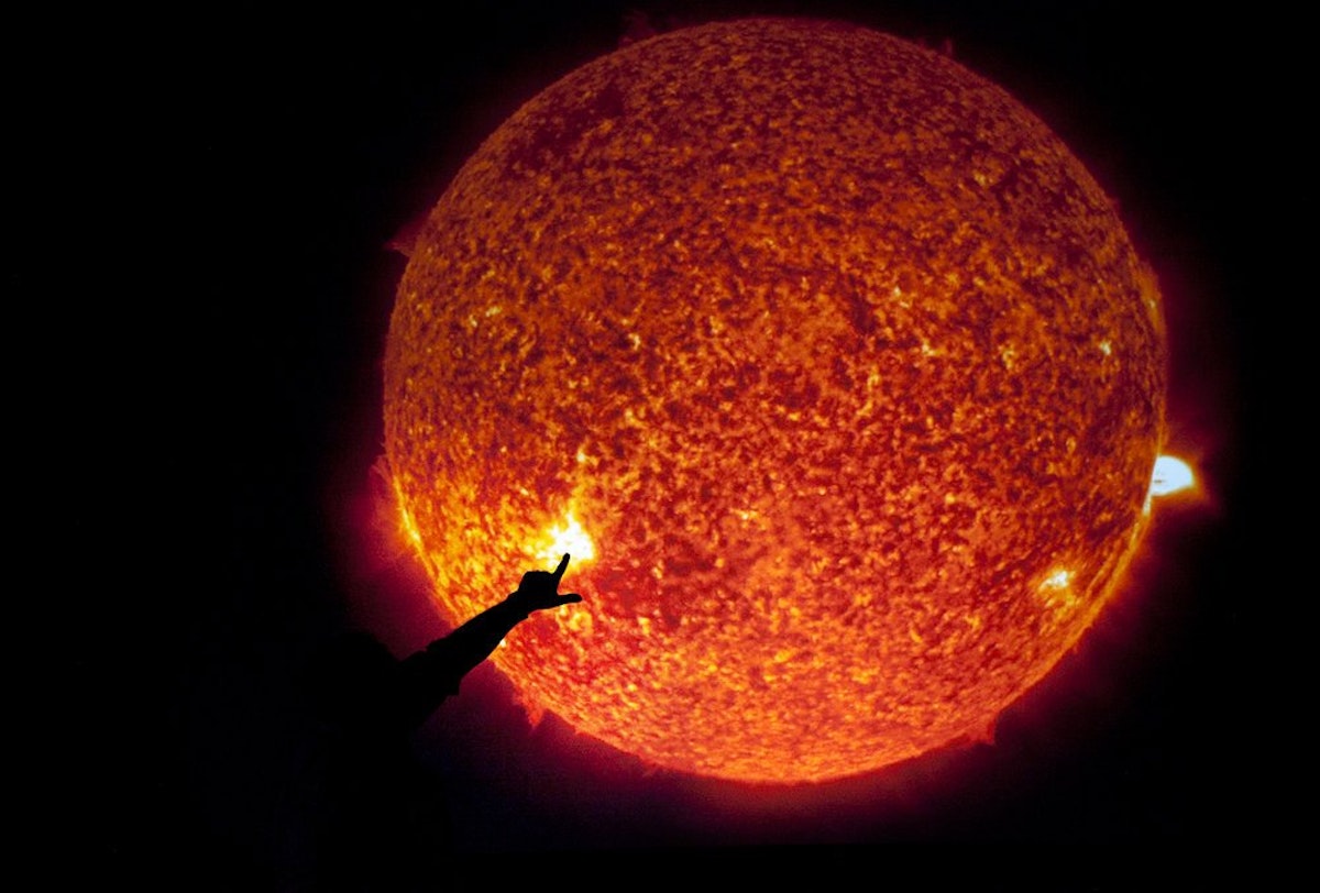 A silhouette of a hand pointing is shown against a clear image of the sun as a burning ball of gas