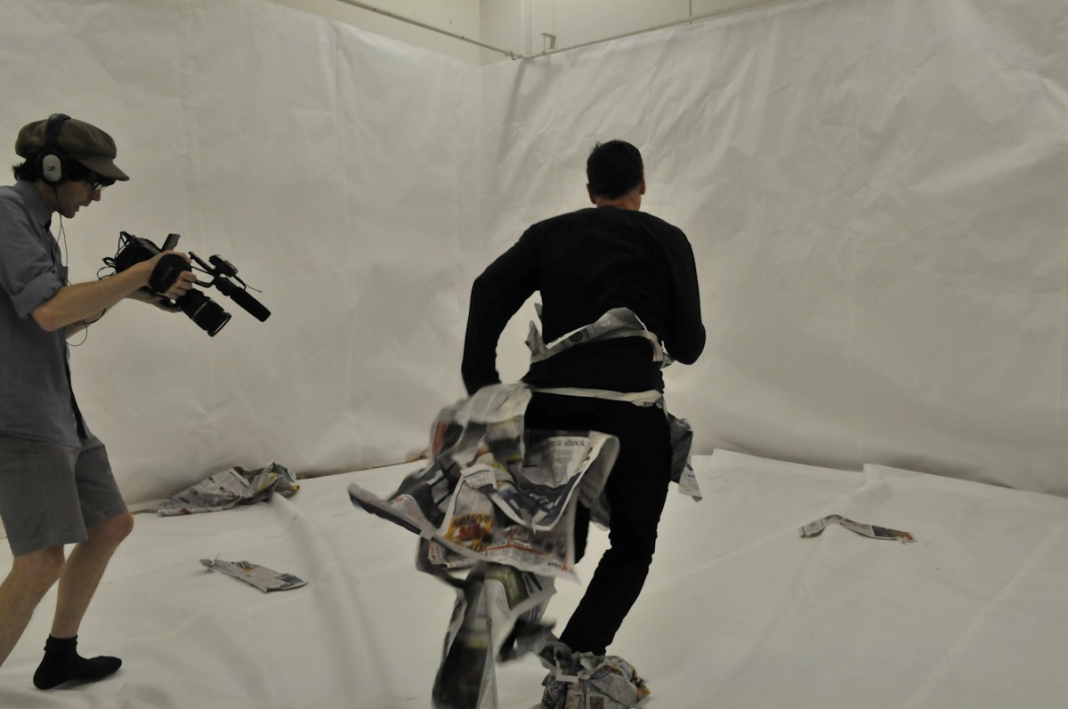 A room lined from floor to ceiling with white paper. At left, a camera operator is shooting a video of the artist Mark Harvey running towards a wall. Mark Harvey is wearing the tattered remains of a costume made entirely of newspaper. The video operator points their camera towards Harvey's feet, which are still covered in newspaper.