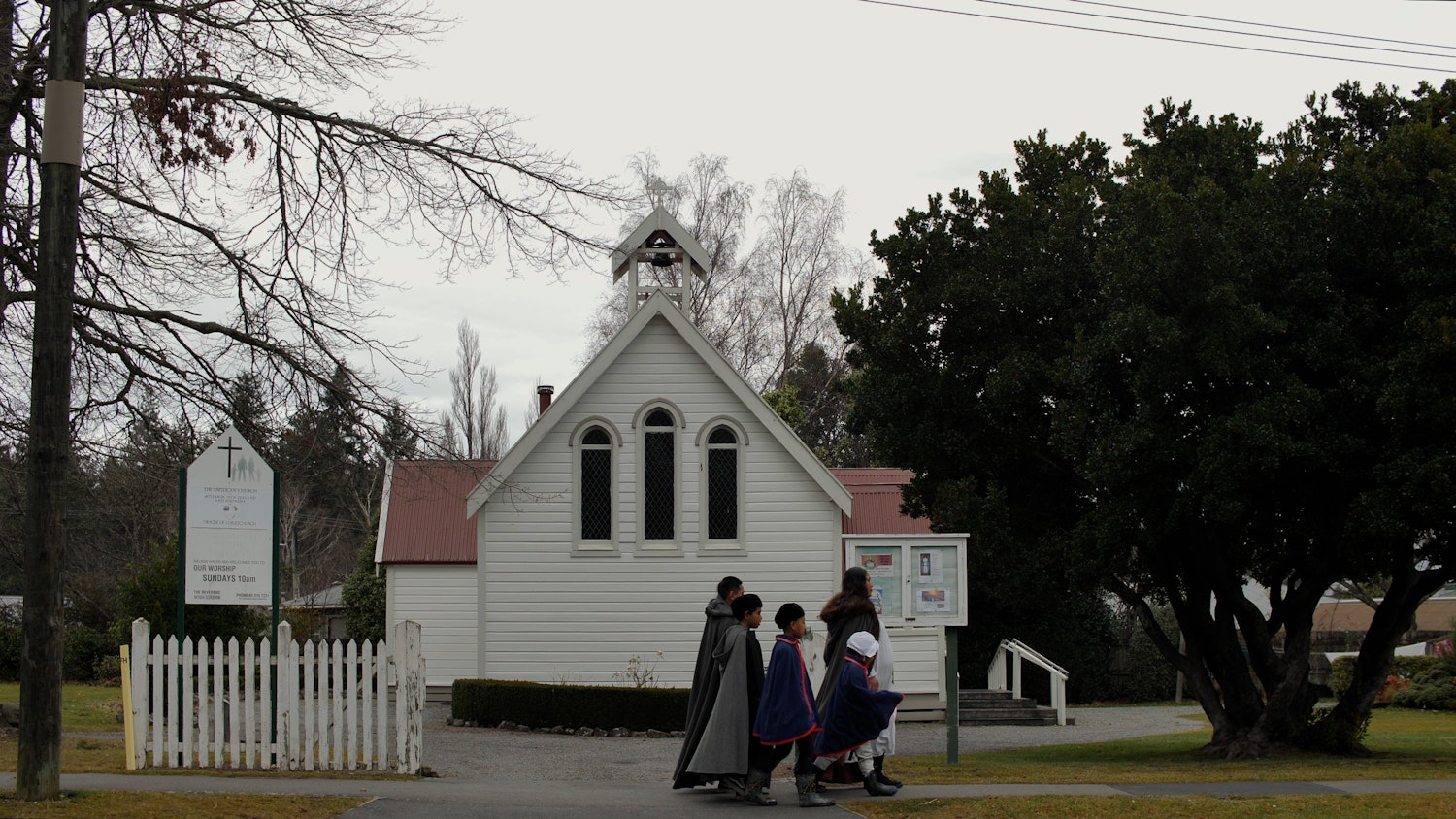 A woman and four children in historic clothing walk past a gothic-seeming wooden church. The sky is grey and ominous.