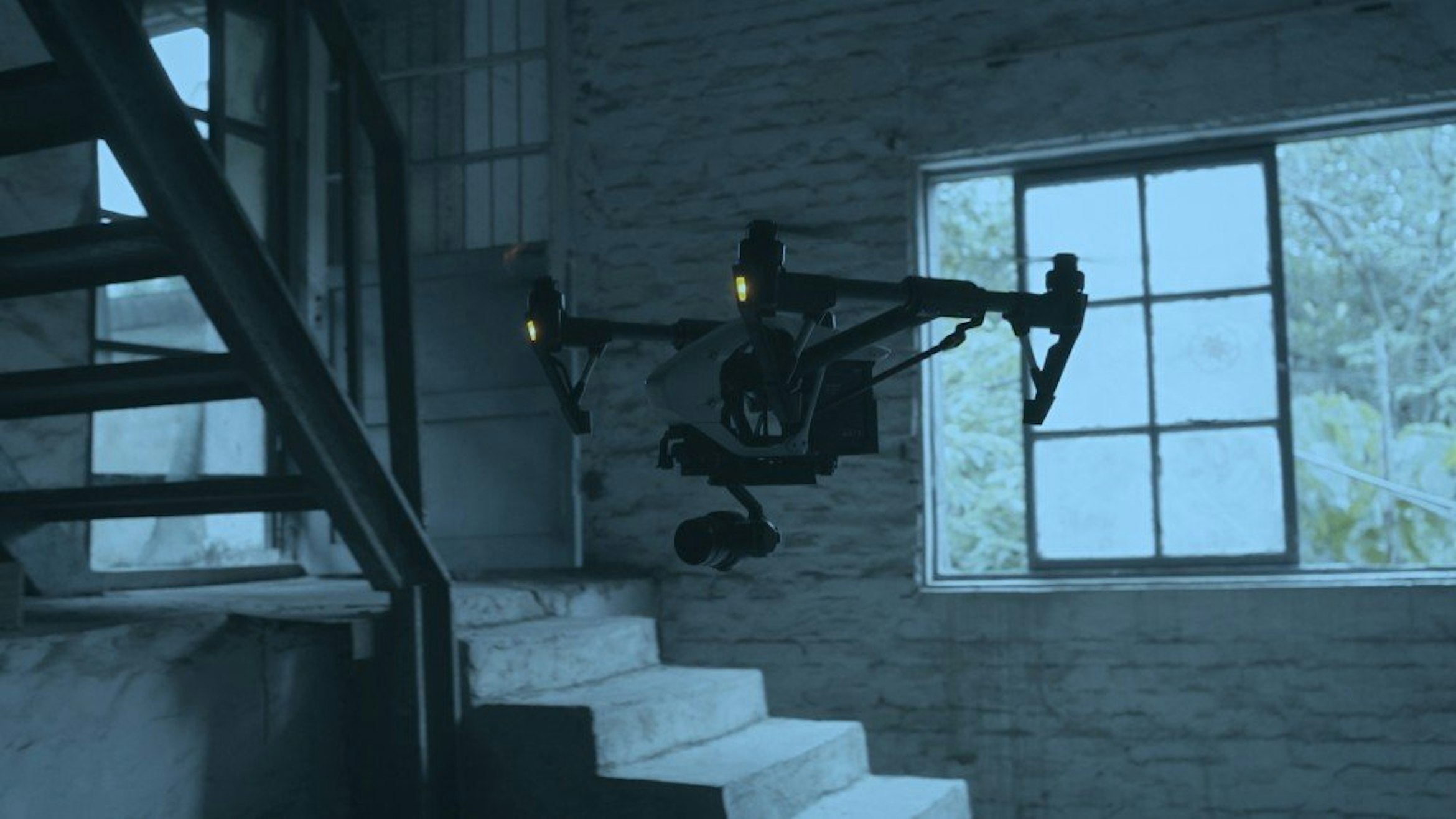 A drone flies through the inside of a building which has concrete steps and a window outside to greenery. The colour of the image is blueish grey as if its dawn, dusk or a very cloudy day