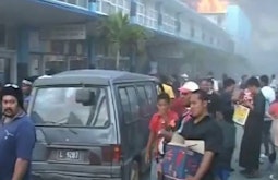A large crowd of people riot on a street in Tonga, there is a building afire in the distance.