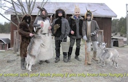 A group of people dressed as goats stand in front of a barn, there are two goats on leads standing with them.