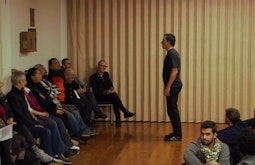 A person performs in front of a small audience of people sitting in a small hall.