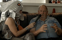 Two people sit on a couch, one is wearing a silver fish costume while interviewing the other person.