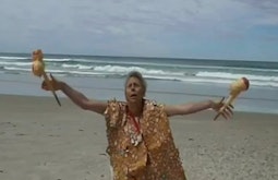A woman does a ritualistic performance on a beach wearing a vest resembling seashells.