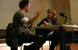 Two people sit at a desk on a stage having a conversation. There is a microphone in front of them.
