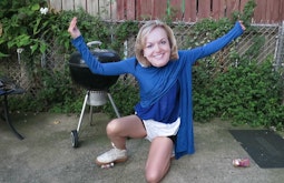 A person wearing blue tops and an oversized Judith Collins mask kneels down in a backyard.