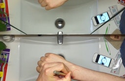At a bathroom sink a person draws a watch-face onto their wrist in red pen.