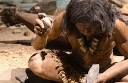 A museum model of an indigenous ancestor sitting down holding a rock in each hand.