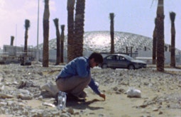A person bends down amongst sand and rocks, there are palm trees and a large dome-topped building behind them.