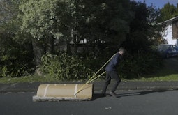 A person drags a roll of cardboard along a road with a yellow strap over their shoulder.