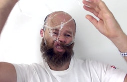 A person wraps sellotape around their face and head, pulling and distorting their features.