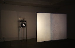 In a gallery an abstract beige image is projected from a slide projector onto a translucent hanging screen.