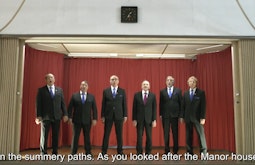 Six men in black suits are singing on a small stage. The subtitles read "on the summery paths. As you looked after the Manor house".