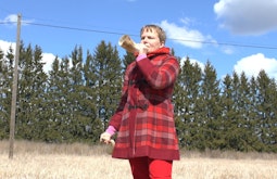 A woman in a red checkered coat stands in a grassy field blowing a horn made from the horn of an animal.