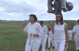 A group of people dressed in white walk through a field holding an inflatable sculpture by strings.
