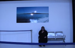 In a dim grey-blue lit room a person sits on an airplane lounge bench looking at their phone. A large photograph of an island is on the wall above them.